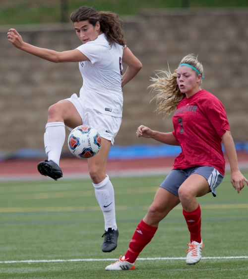 A woman in white in the foreground in midair about to kick a soccer ball while a defensive player readies herself. 
